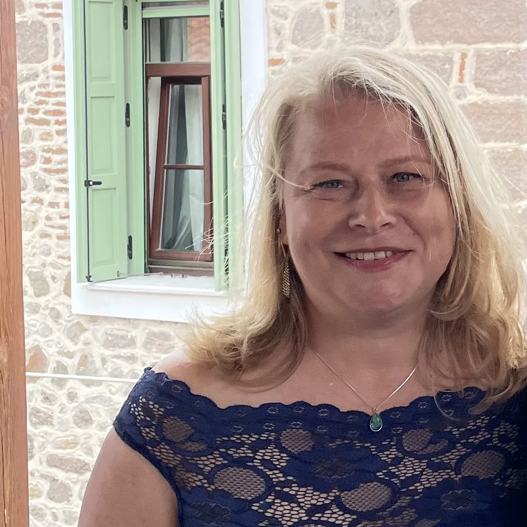 Hanna smiling on a balcony, in front of a stone house with green shutters.