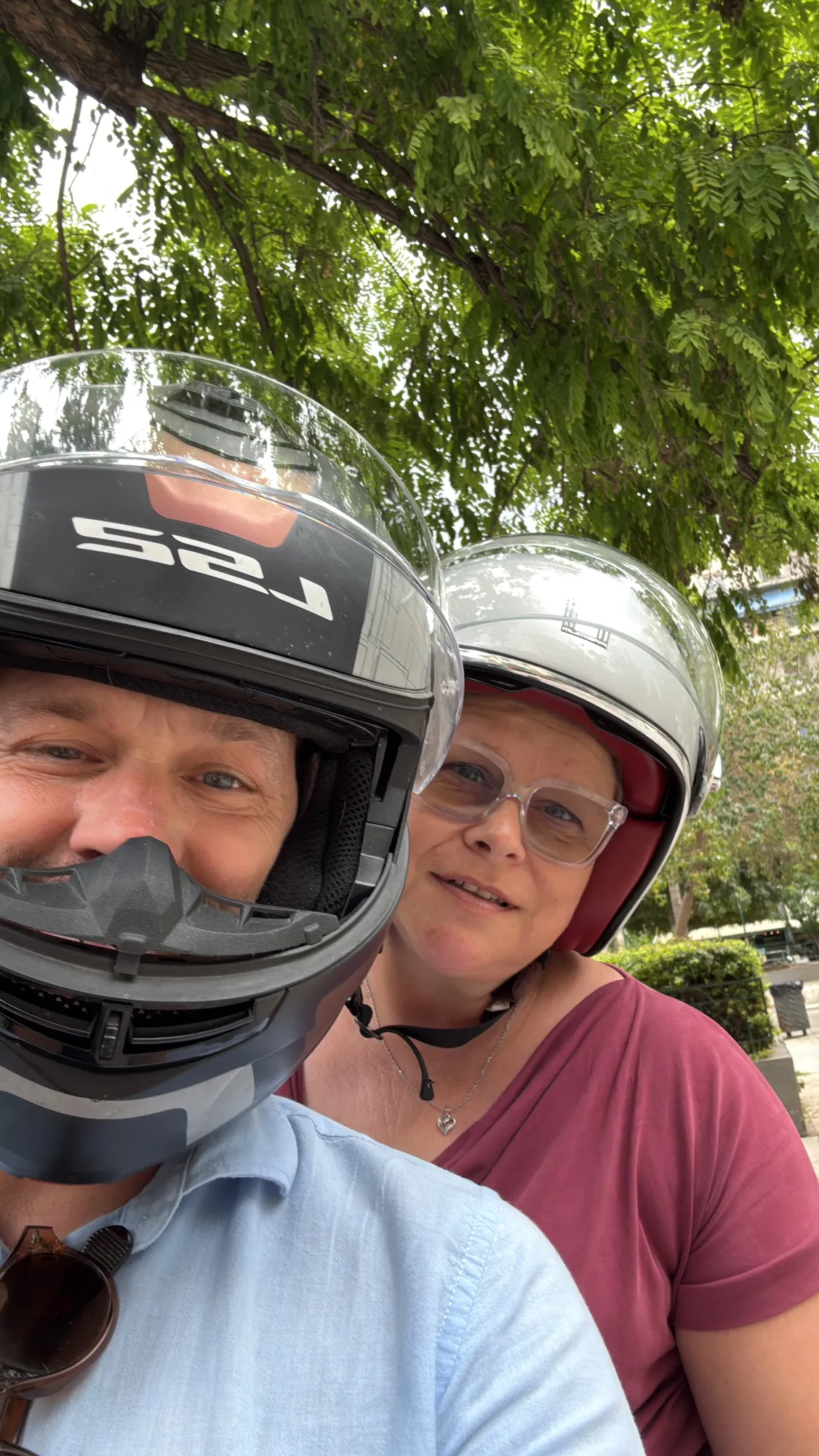 Two people wearing motorcycle helmets posing together outdoors under tree branches.