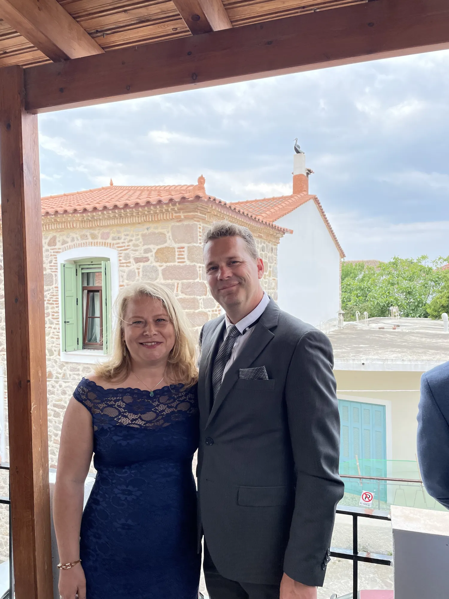 Two people posing together on a covered porch with historic stone buildings visible in the background.