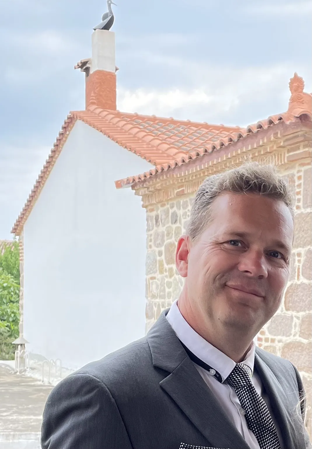 A man in a dark suit smiling in front of a brick building with terracotta roof tiles.